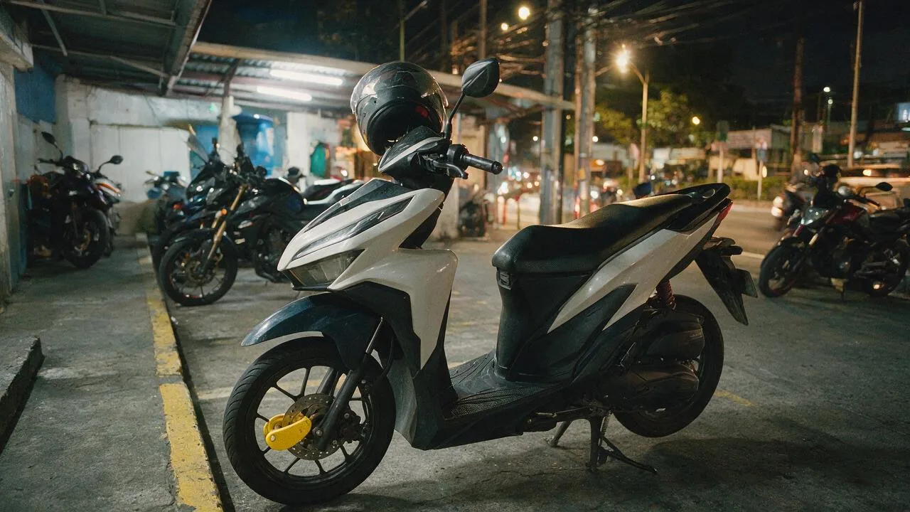 A scooter parked at night with a yellow disc lock on the front wheel and a helmet on the mirror